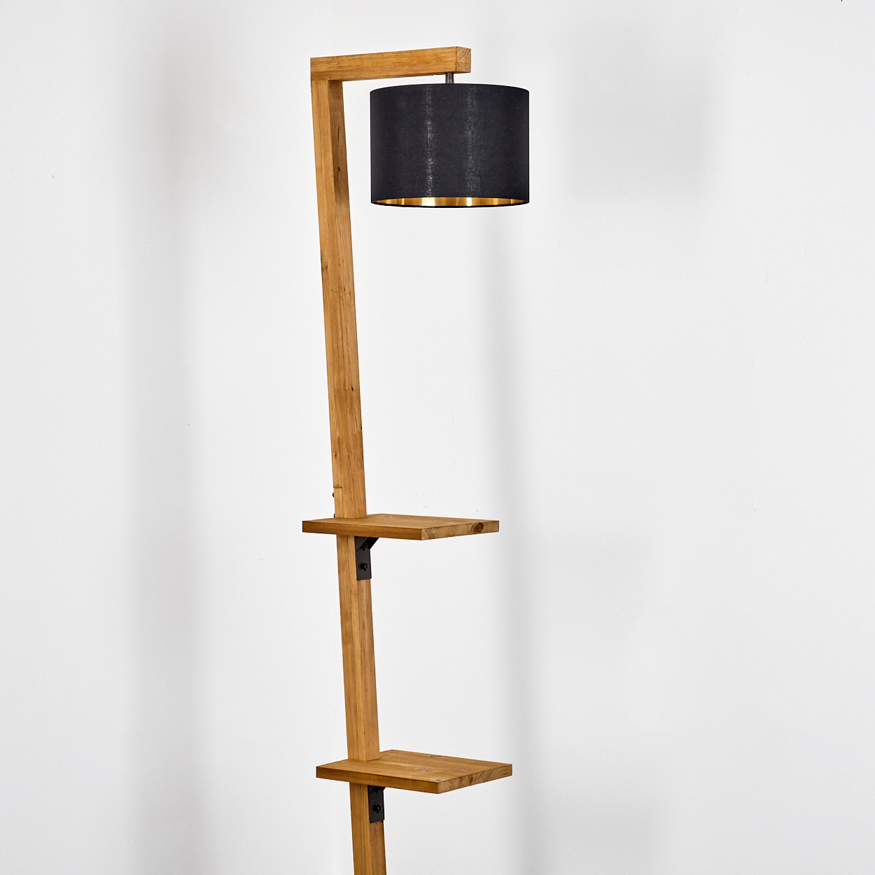 Eine hohe Holz-Stehlampe mit schwarzem Lampenschirm. Sie hat zwei Regale und steht vor einer weißen Wand.
