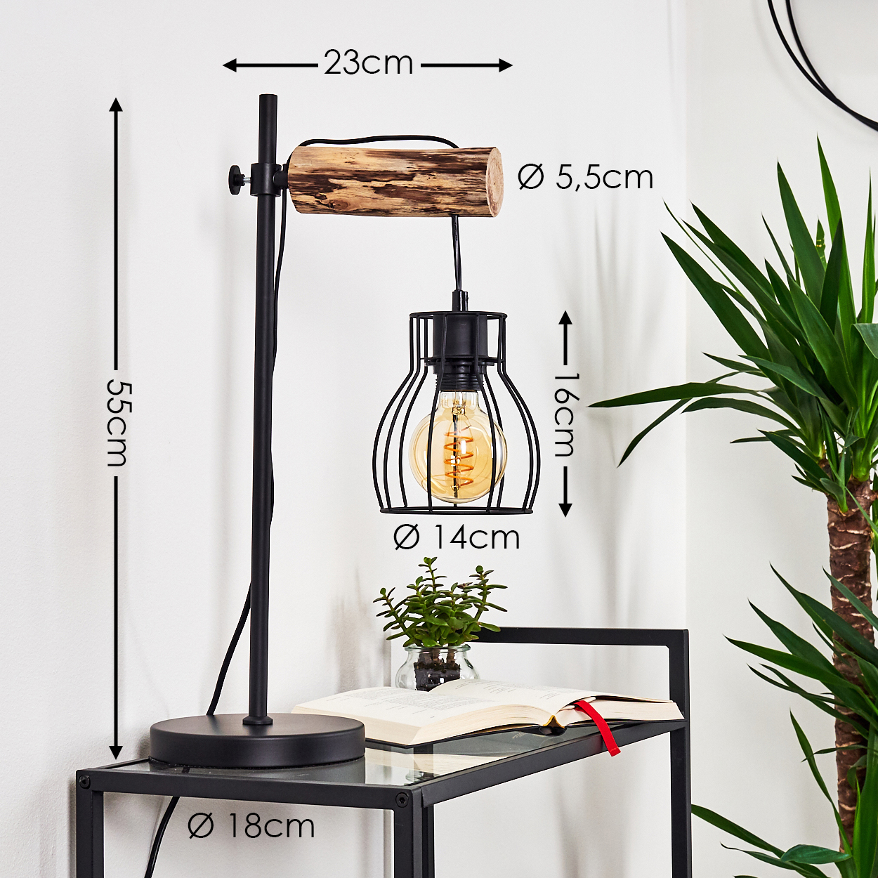 Eine schwarze Lampe mit Holzarm steht auf einem Tisch. Ein Buch und eine Pflanze stehen ebenfalls auf dem Tisch.