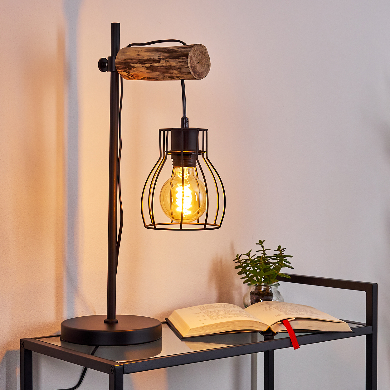 Eine schwarze Lampe mit Holzaufsatz, einem vergitterten Leuchtmittel und ein Buch auf Glastisch.