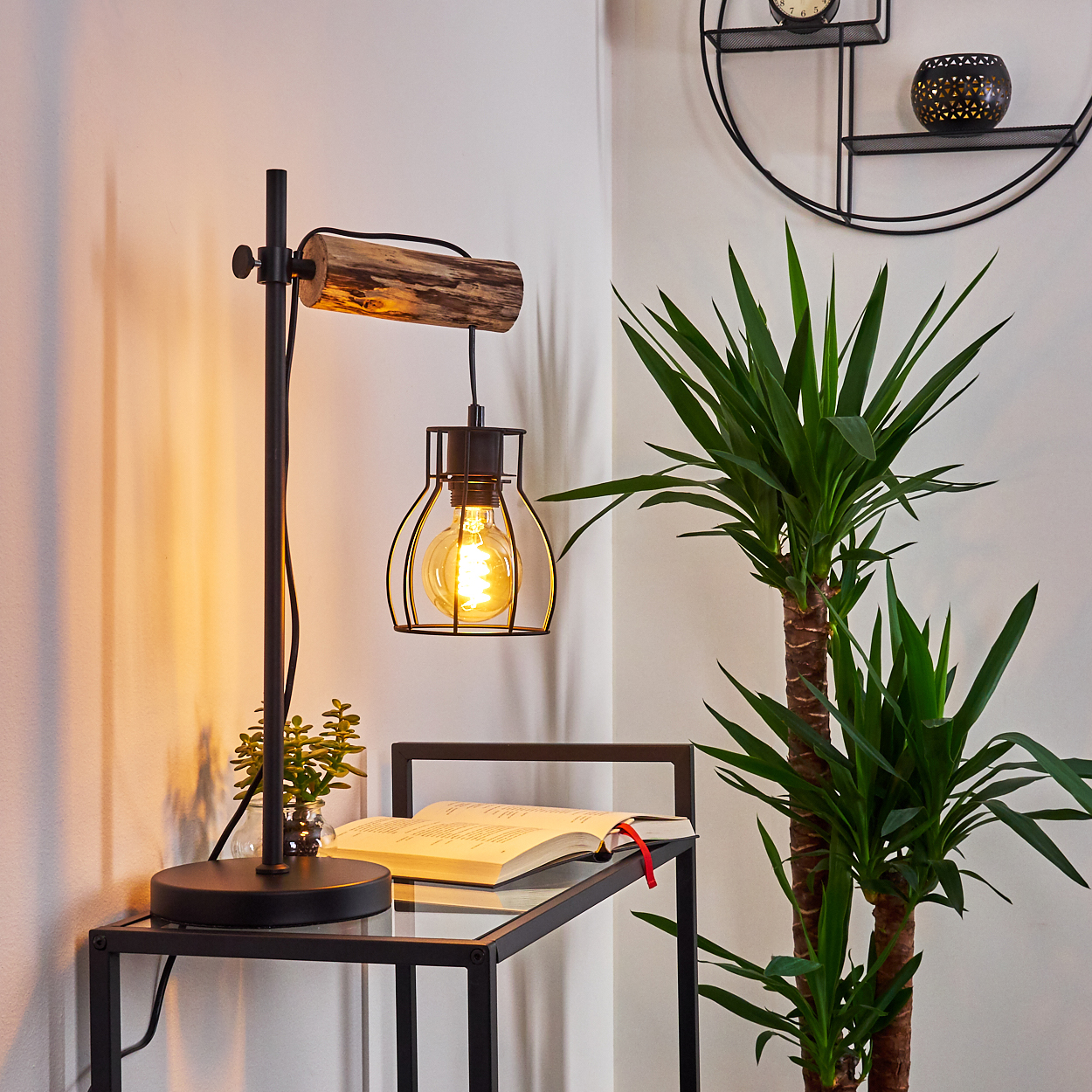 Eine schwarze Lampe mit Holzarm. Ein Buch und eine kleine Pflanze stehen auf dem Tisch. Eine große Pflanze steht daneben.