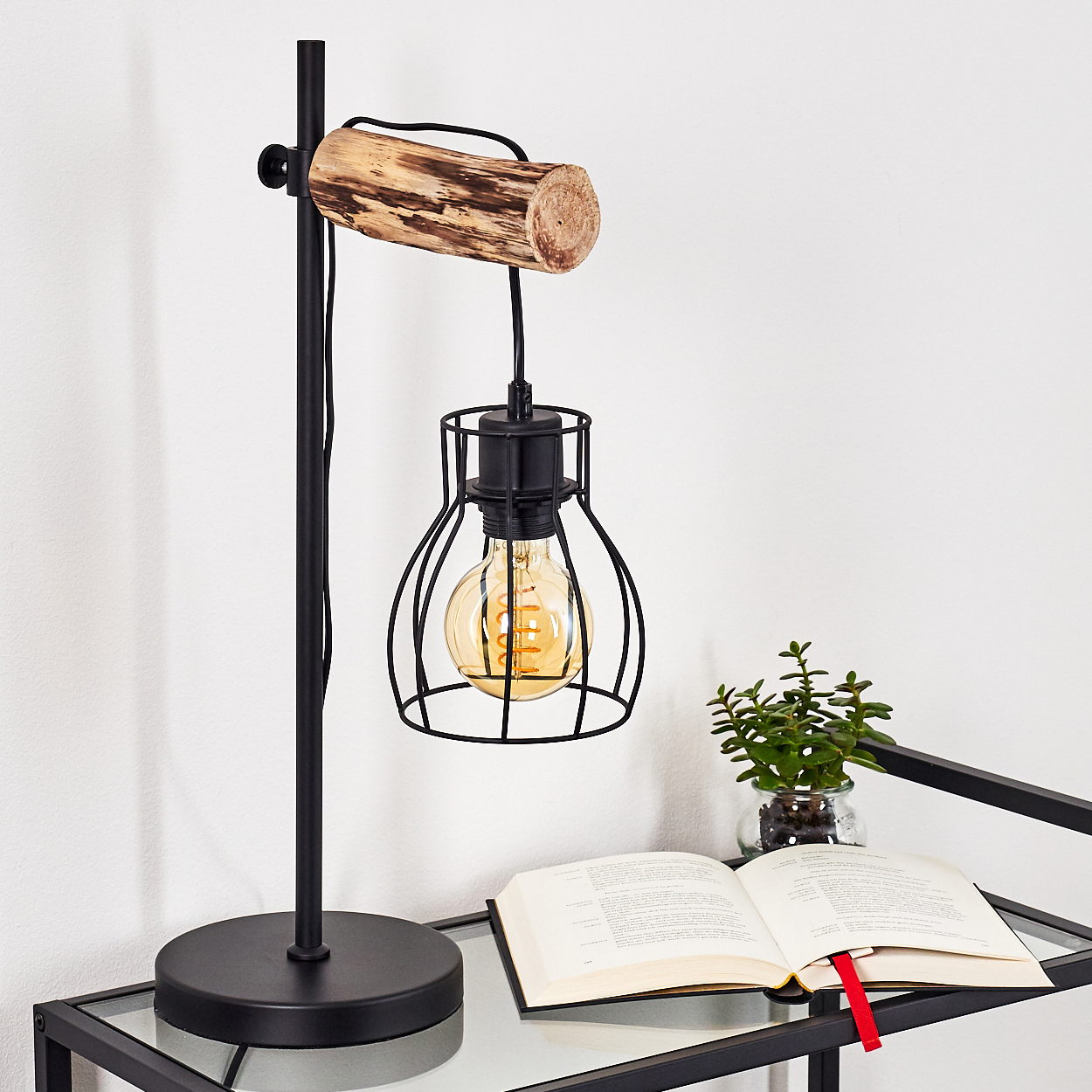 Eine schwarze Lampe mit Holzarm. Ein Buch und eine kleine Pflanze stehen auf einem Glastisch.