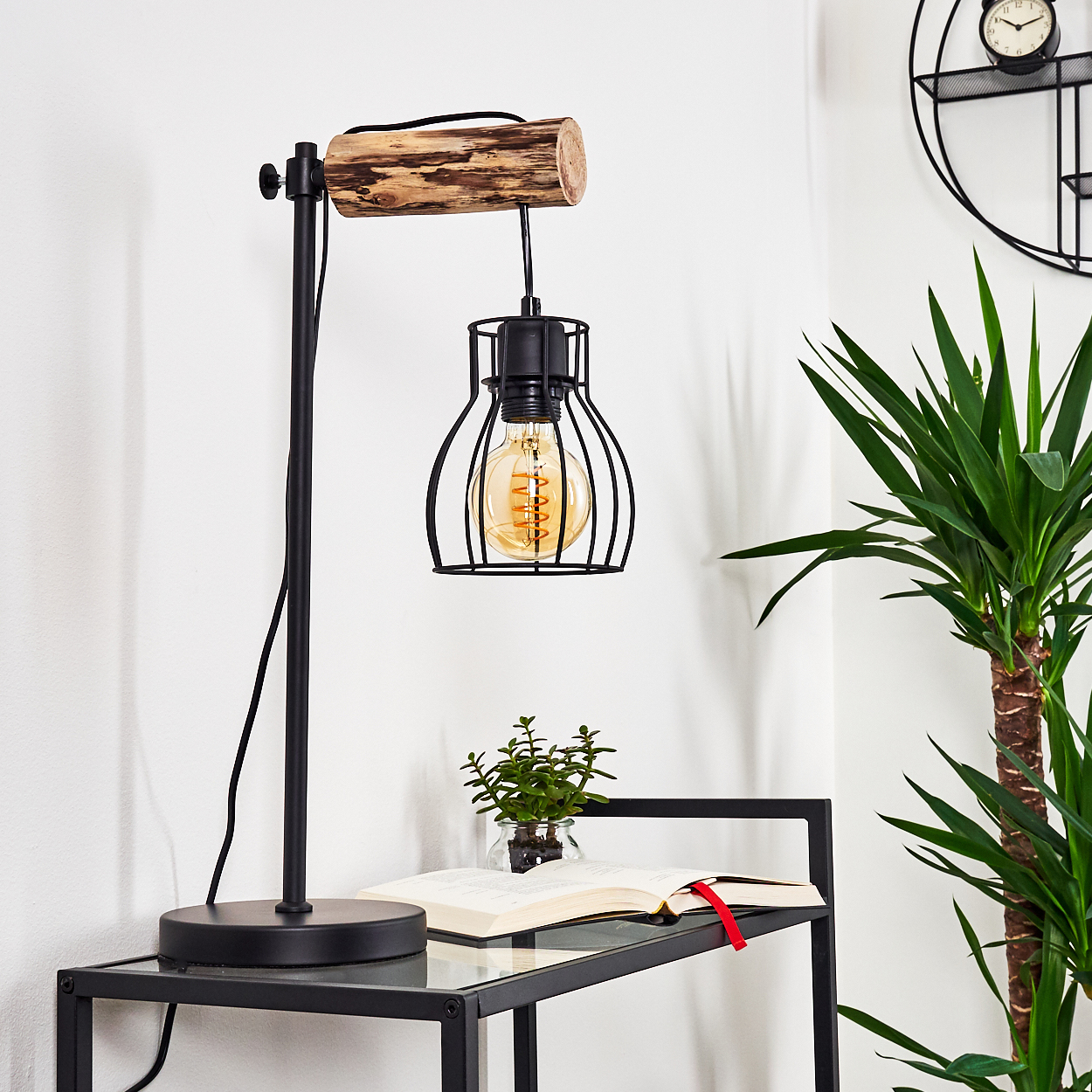 Eine schwarze Schreibtischlampe mit Holzbalken, die eine Käfigbirne hält, auf einem Glastisch mit Buch und Pflanze.