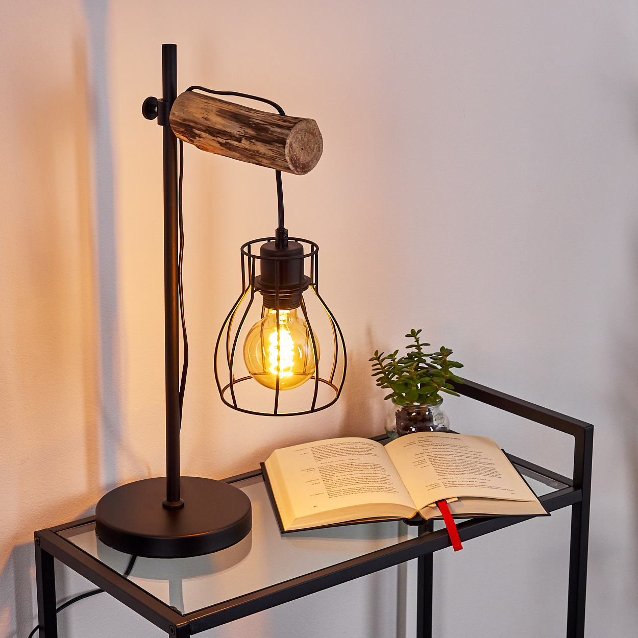 Eine leuchtende Schreibtischlampe mit Käfigbirne, einem Buch und einer kleinen Pflanze auf einem Glastisch. Weiße Wand im Hintergrund.