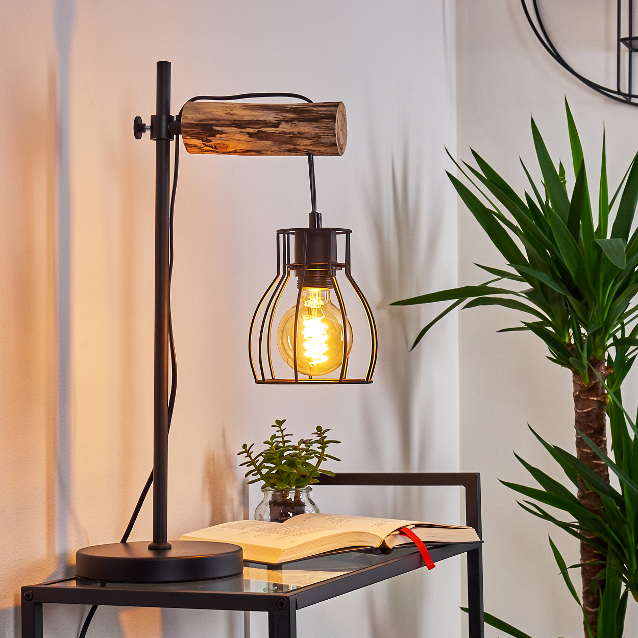 Schreibtischlampe mit Käfigbirne, Buch und Pflanze auf Glastisch. Weiße Wand mit dekorativer Uhr und Pflanze.