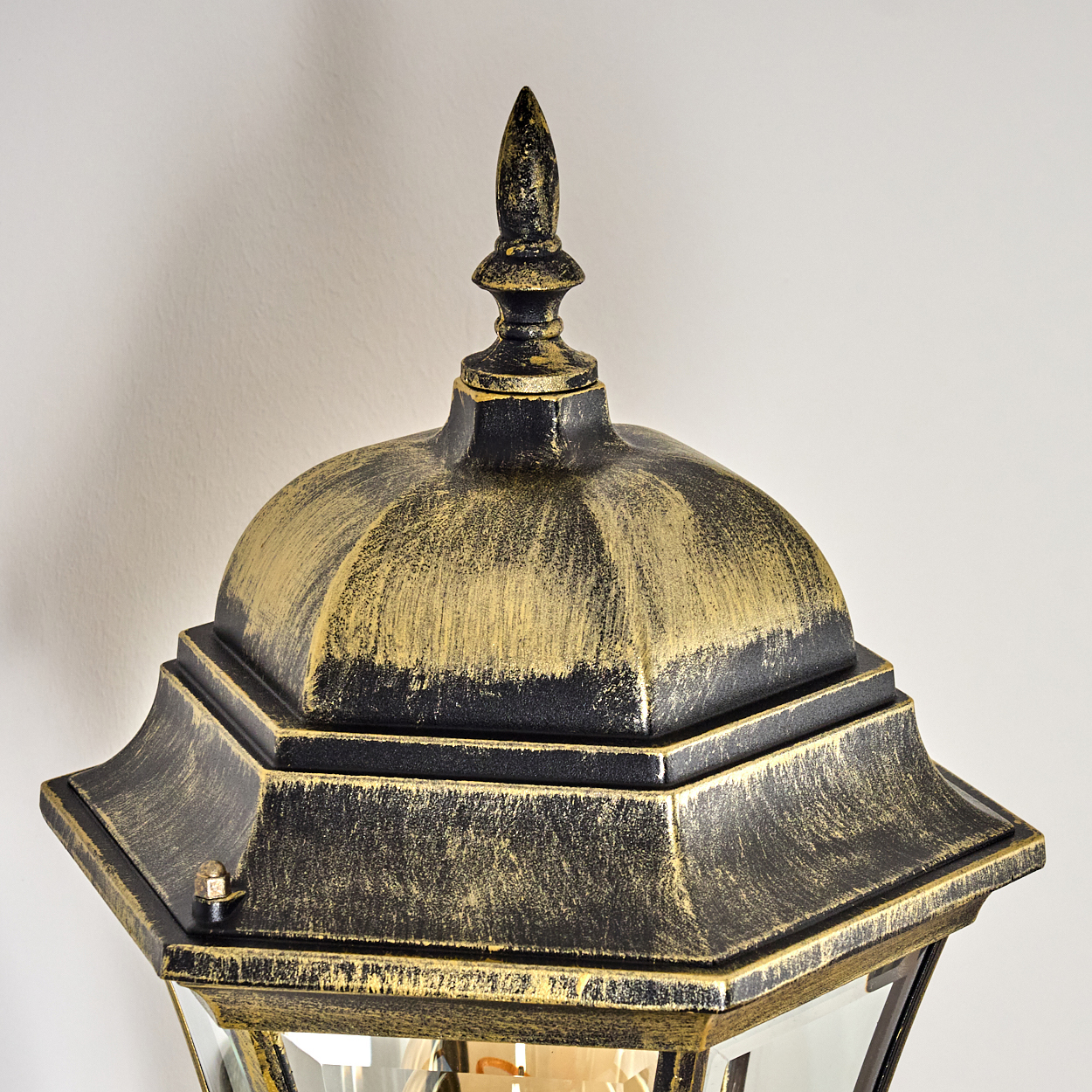 Nahaufnahme der Oberseite einer schwarz-goldenen Wandleuchte. Die Lampe hat eine Glasfront und ist an einer weißen Wand montiert.