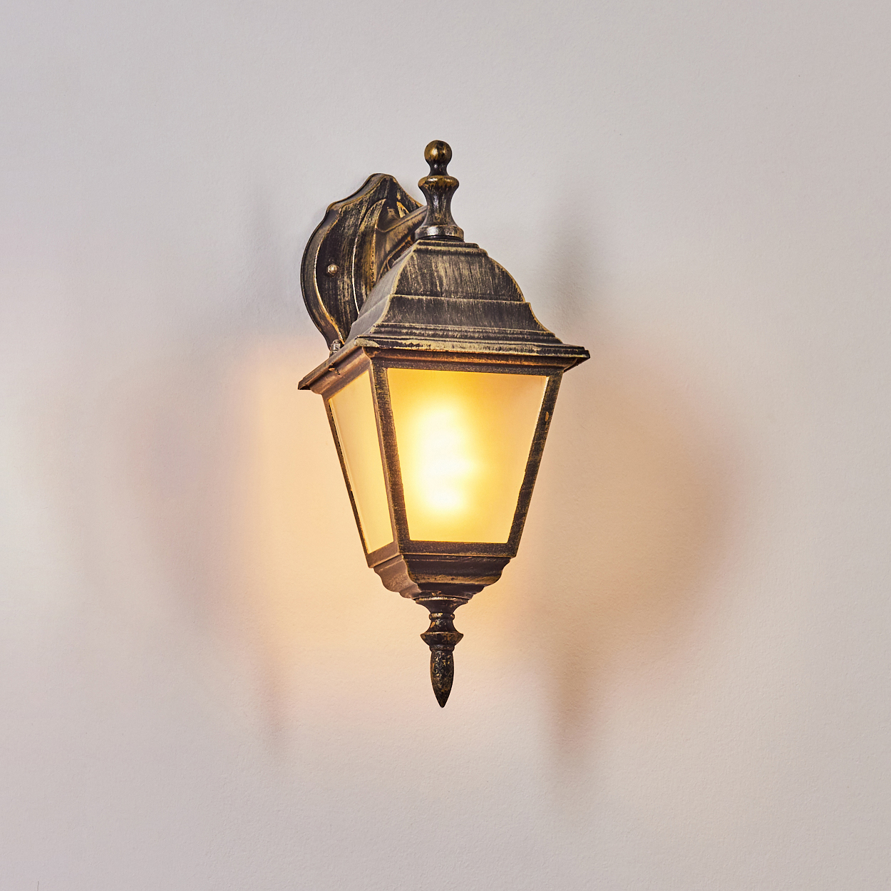 Eine schwarz-goldene Wandleuchte mit leuchtender Glühbirne. Die Lampe hat eine Glasfront und ist an einer weißen Wand montiert.