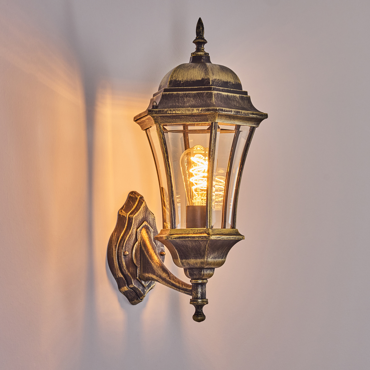 Eine schwarz-goldene Wandleuchte mit leuchtender Glühbirne. Die Lampe hat eine Glasfront und ist an einer weißen Wand montiert.