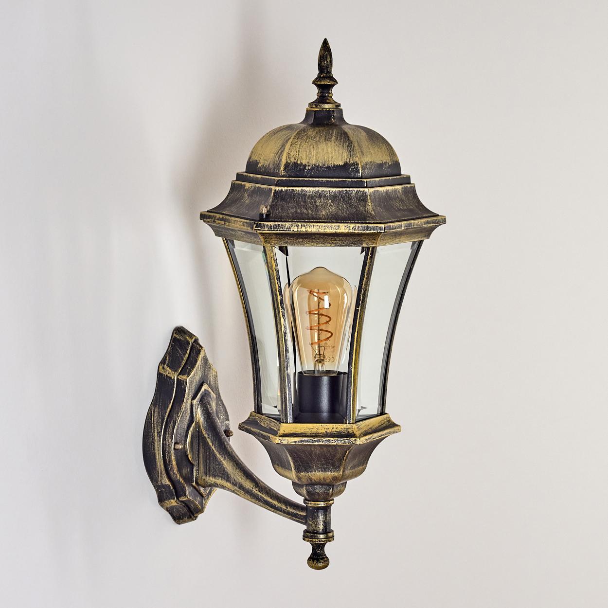 Eine antike goldene Wandlampe mit klarem Glasschirm und Wendelglühbirne, an einer weißen Wand befestigt.