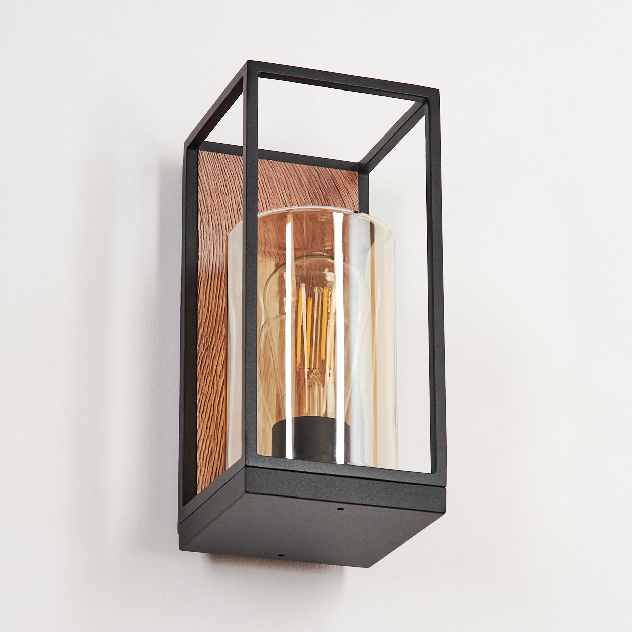 Eine schwarze, quadratische Wandleuchte mit Holzrückwand und klarem Glaszylinder, beleuchtet von einer Wendelglühbirne.