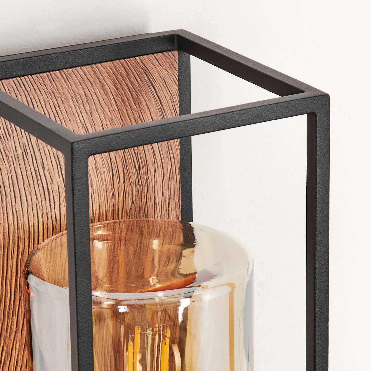 Nahaufnahme einer Wandleuchte mit Holzrücken, schwarzem Rahmen und einem zylindrischen Glasschirm mit Glühbirne.
