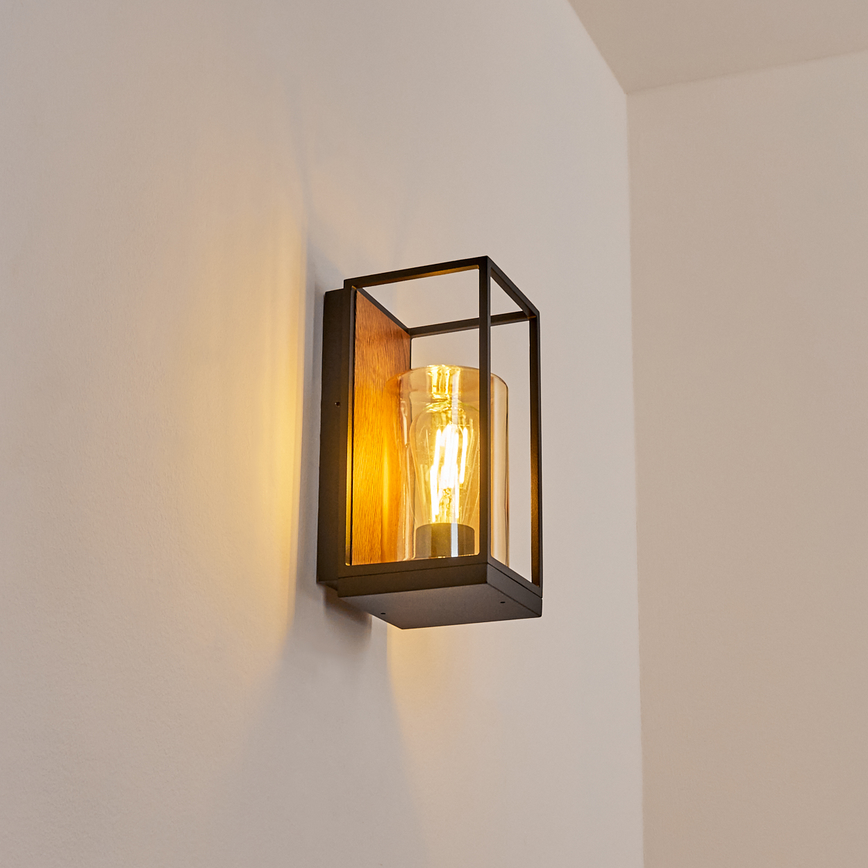 Eine Wandleuchte mit schwarzem Rahmen, Holzrückseite und einem Gehäuse aus klarem Glas. Die Lampe befindet sich an einer weißen Wand.