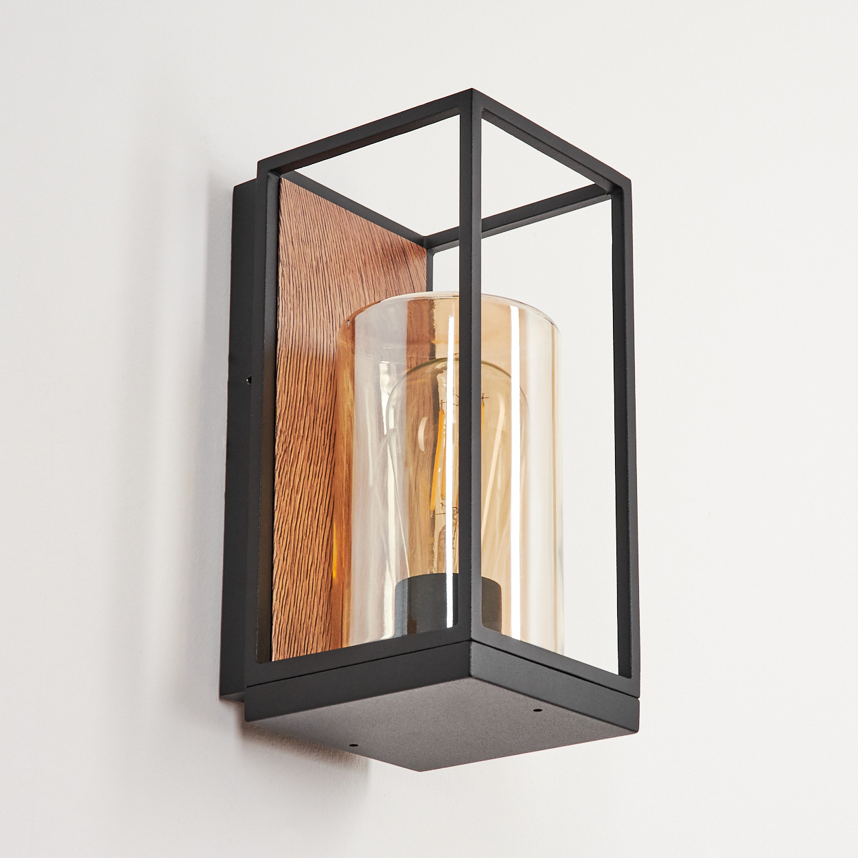 Eine Wandlampe mit schwarzem Rahmen, Holzrücken, zylindrischem Glasschirm und Glühbirne, an einer weißen Wand montiert.