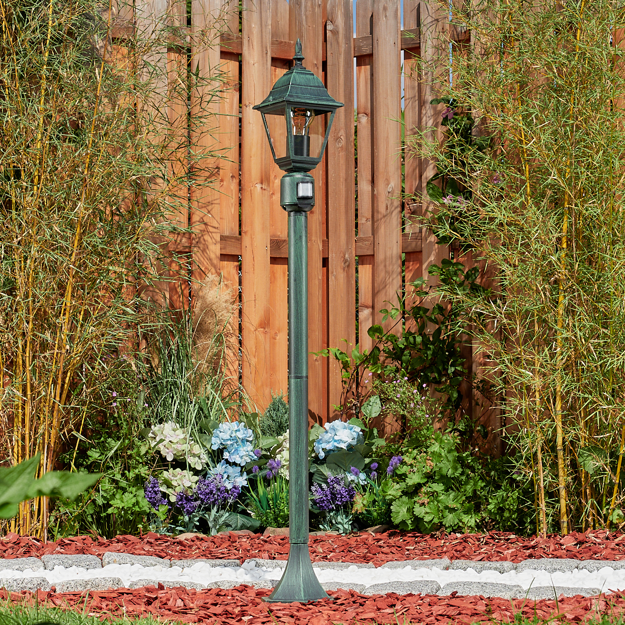 Grüne Außenlaterne im Garten. Mit Licht, umgeben von Pflanzen und einem Zaun.