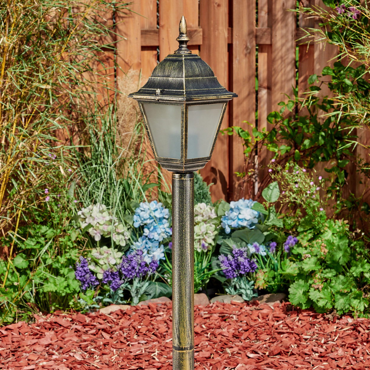 Außenlaterne im Garten. Mit Licht, zwischen Pflanzen und roter Mulch.