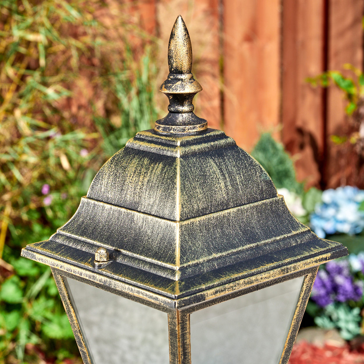 Nahaufnahme des oberen Teils einer gold-schwarzen Lampe. Der Hintergrund ist ein Holzzaun und grüne Pflanzen.