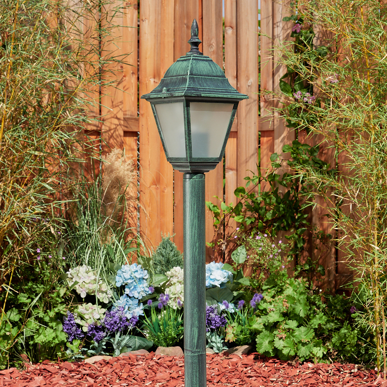 Ein grüner Lampenpfahl in einem Garten, mit einem Holzzaun und grünen Pflanzen im Hintergrund.