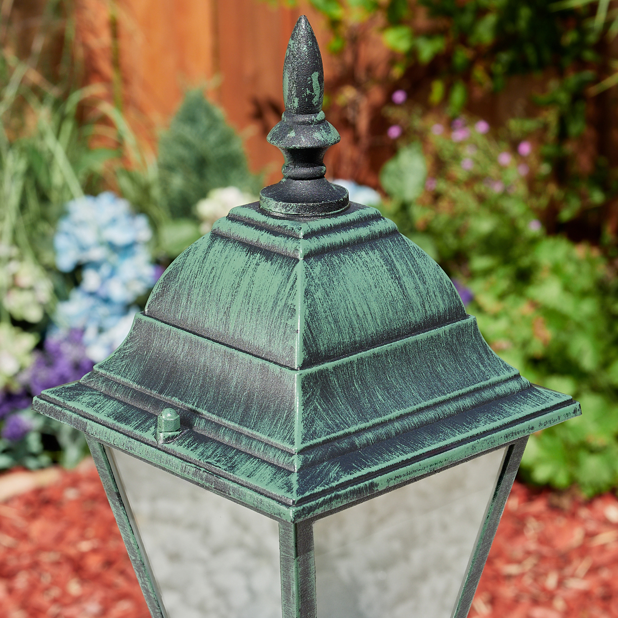 Nahaufnahme des oberen Teils einer grün-schwarzen Außenlampe mit spitzer Spitze, vor einem verschwommenen Gartenhintergrund.