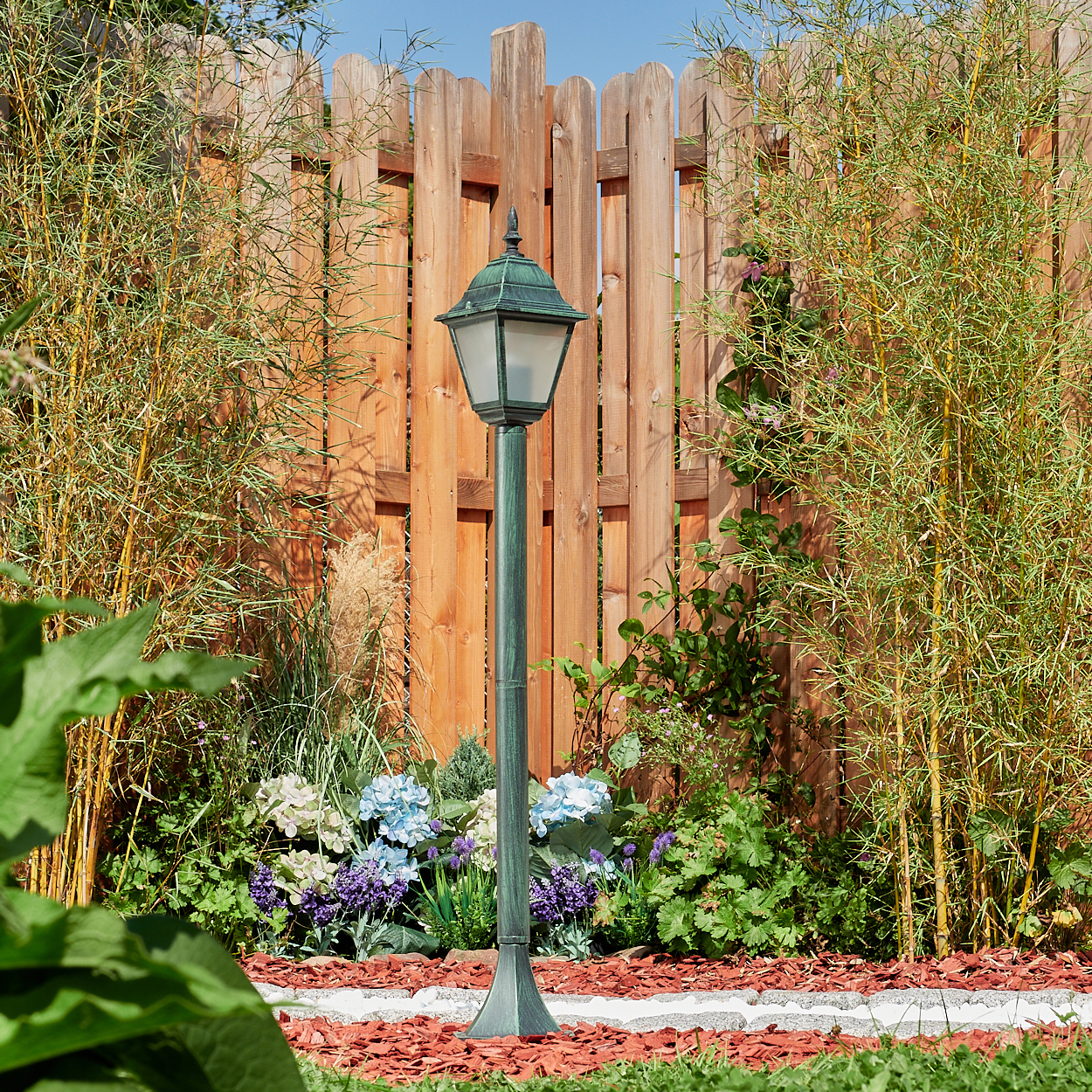 Laternenpfahl in einer Gartenszene mit Holzzaun und Bambus. Die Lampe hat eine grün-schwarze Oberfläche.