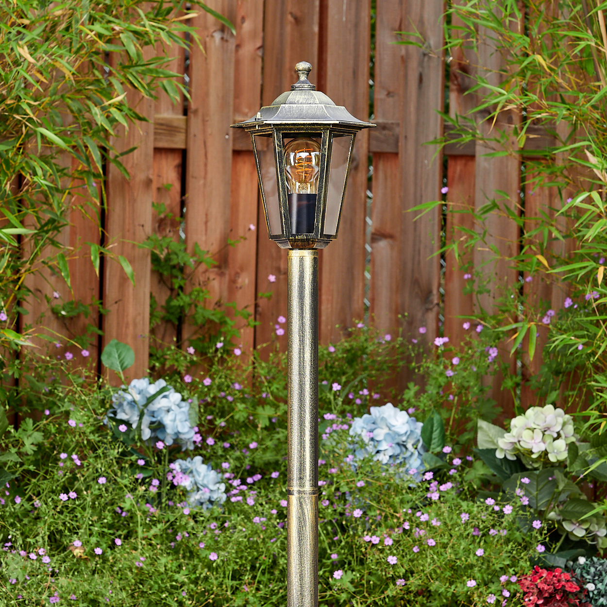 Eine Laterne in einem Garten, mit Licht, umgeben von Blumen und einem Holzzaun.