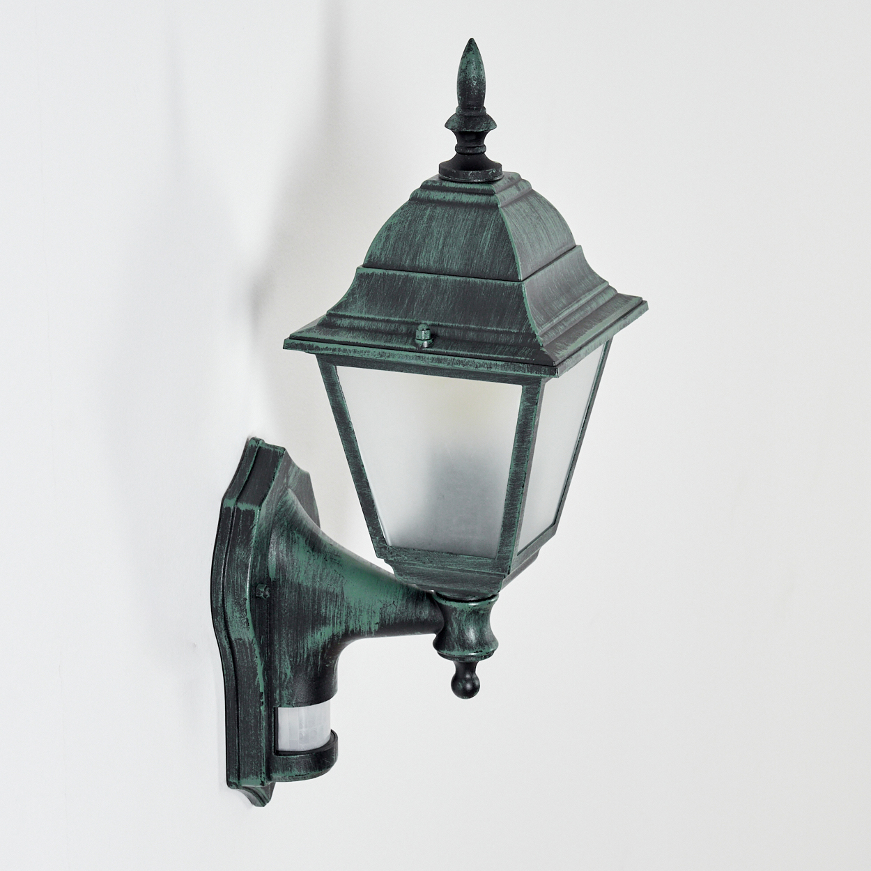 Grüne Außenlampe mit Licht und Bewegungsmelder an einer weißen Wand.