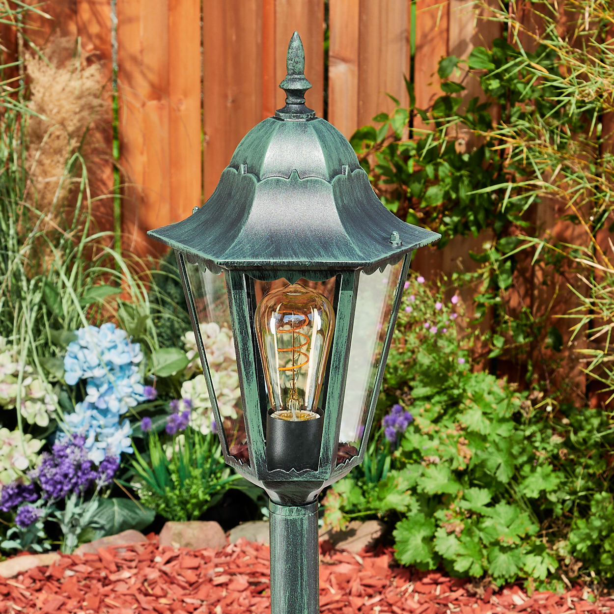 Grüne Außenlampe mit dekorativer Spitze, Glasscheiben und einer leuchtenden Glühbirne im Garten.