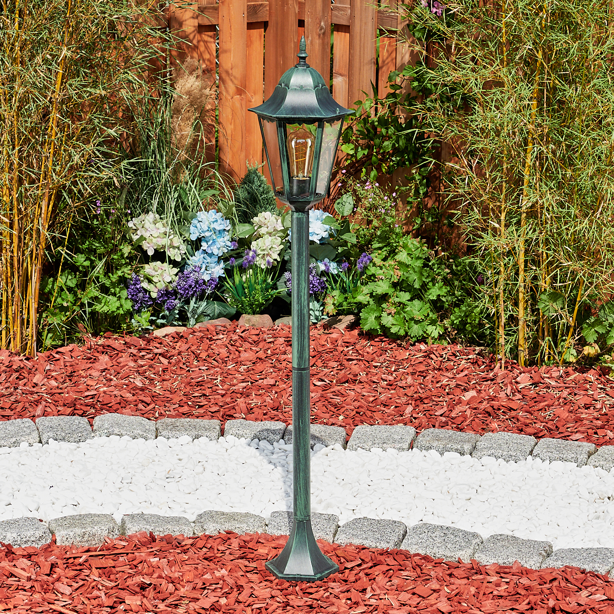 Hohe, grüne Außenlampe mit Glühbirne. In einem Garten mit roter Mulch und Bambus aufgestellt.
