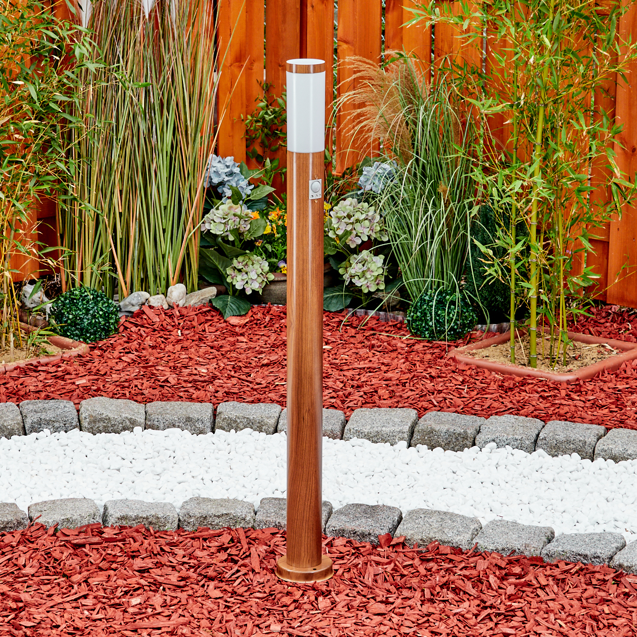 Hohe Holz-Außenlampe, umgeben von roter Mulch und Pflanzen, in der Nähe eines Holzzauns.