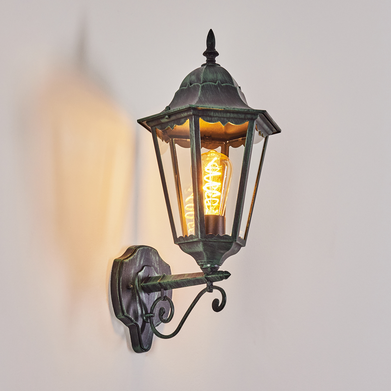 Eine Wandlampe für den Außenbereich mit grünem Metallic-Finish und sichtbarer Glühbirne. Hintergrund ist eine weiße Wand.