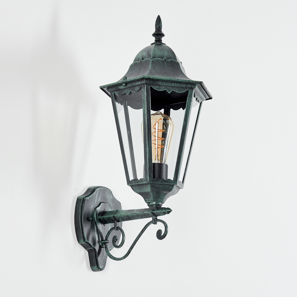 Eine Außenwandleuchte mit grün getöntem, verziertem Metallrahmen, klarem Glas und einer leuchtenden, warmtonigen Glühbirne.