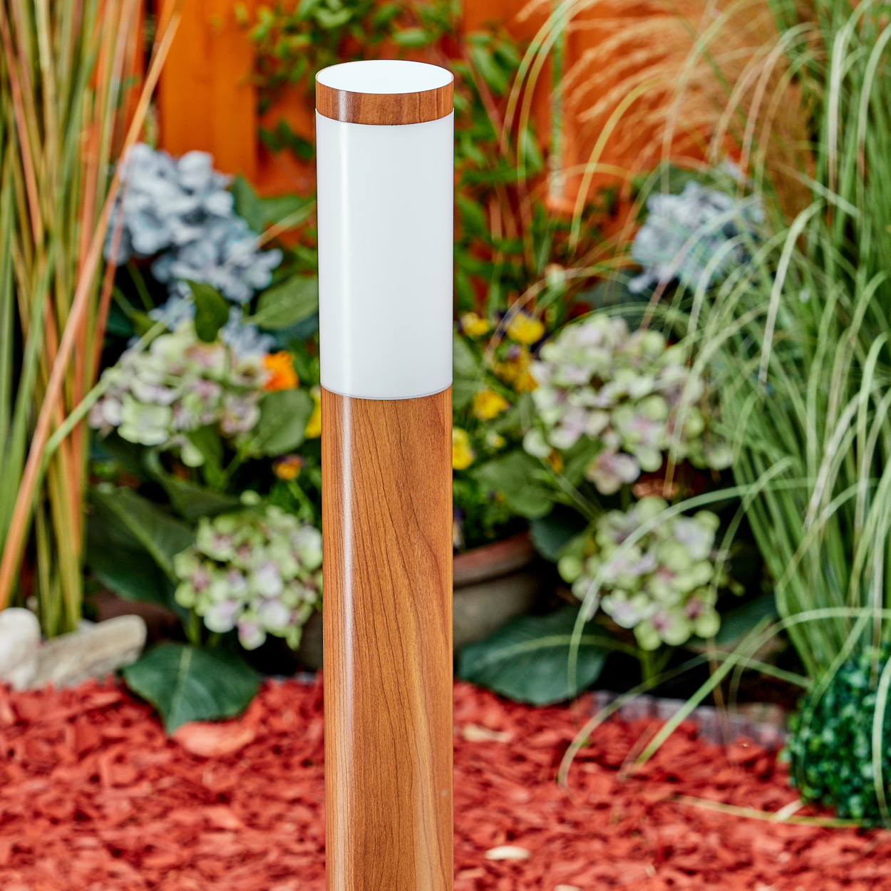 Eine hohe, holzartig aussehende Polleuchte mit weißer Spitze steht in einem Garten mit roter Mulch und Grün.
