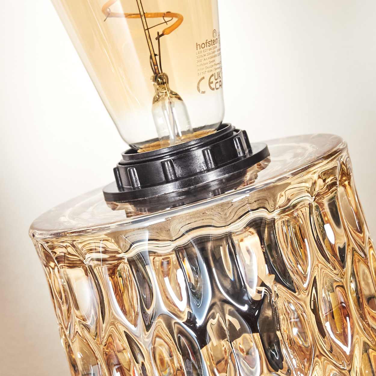 Nahaufnahme einer Lampe mit strukturiertem bernsteinfarbenem Glasfuß und einer leuchtenden Glühbirne. Der Hintergrund ist unscharf.