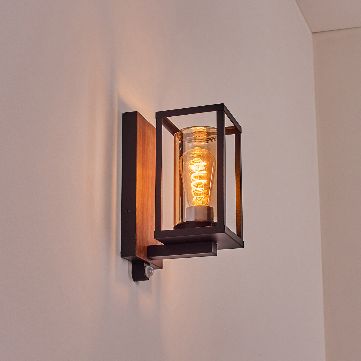 Eine Wandlampe mit schwarzem Rahmen, Holzhintergrund und sichtbarer Glühbirne, an einer weißen Wand befestigt.