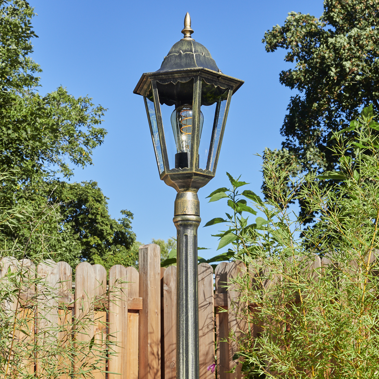 Außenlaterne mit dekorativer Oberseite, in einem Garten mit Holzzaun und Grün.