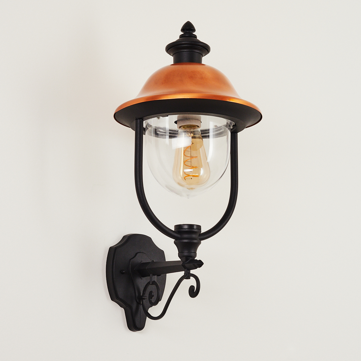 Eine Außenwandlampe mit kupferfarbenem Aufsatz, schwarzem Rahmen und klarem Glas. Sie ist an einer weißen Wand montiert.