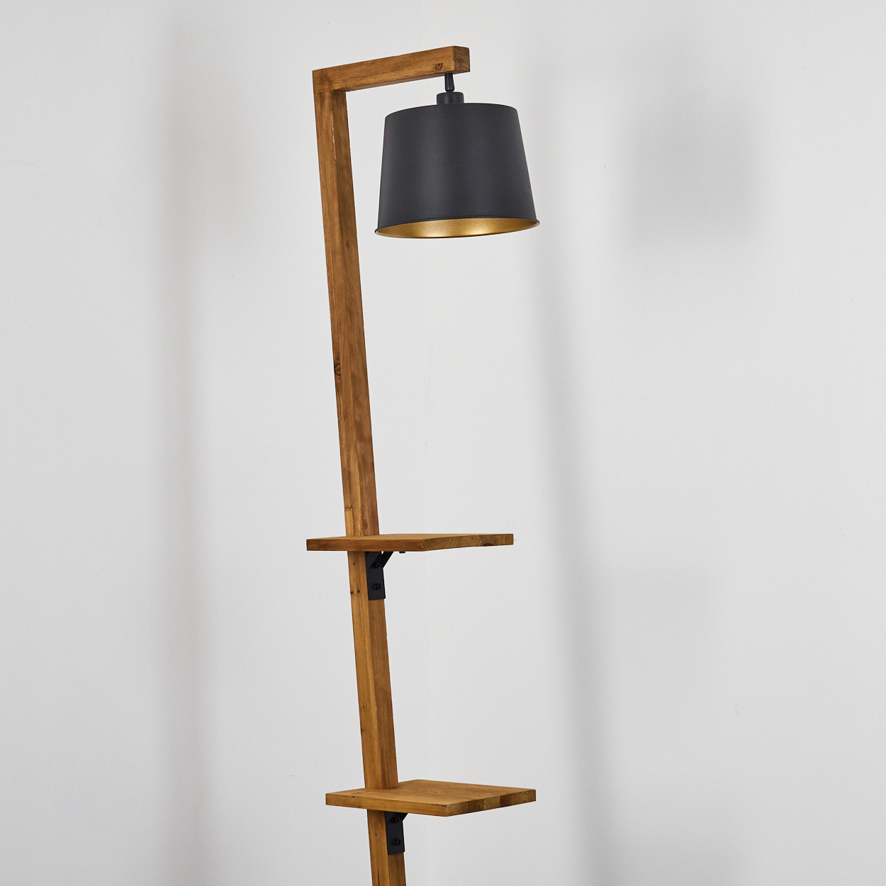 Eine hohe Holzlampe mit zwei Regalen und einem schwarzen Lampenschirm. Die Lampe steht vor einer weißen Wand.