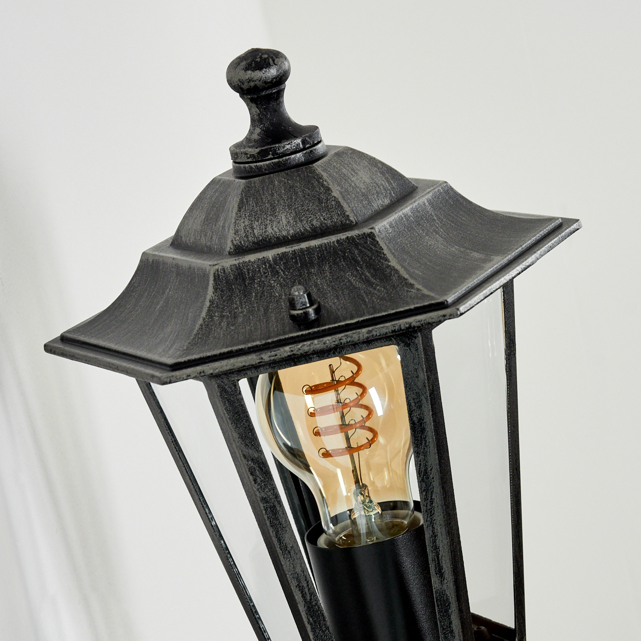 Nahaufnahme der Oberseite einer schwarzen und silbernen Außenlampe mit Glühbirne, schräg vor weißer Wand.