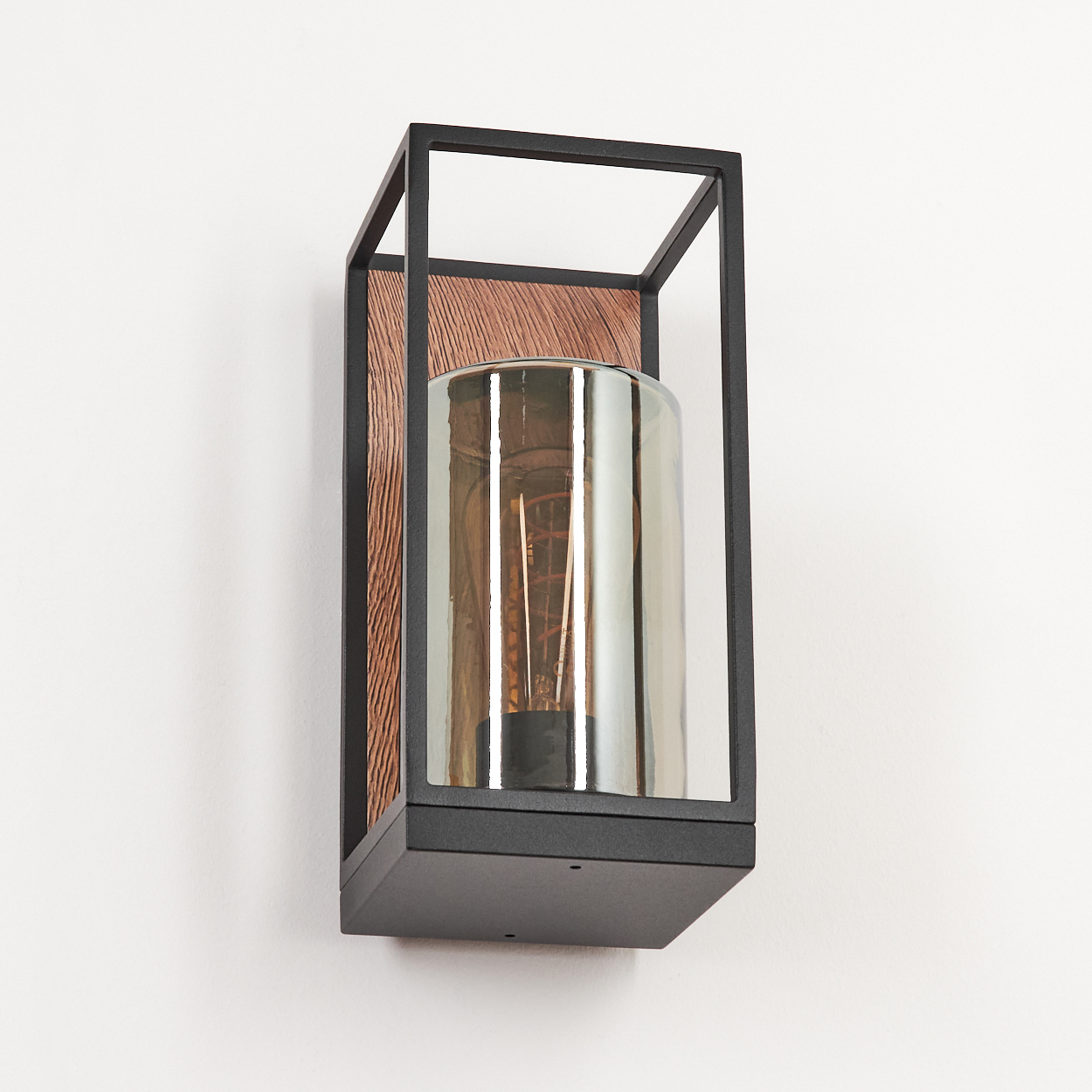 Eine moderne Wandleuchte mit schwarzem Rahmen, Holzhintergrund und zylindrischem Glasschirm. Sie ist an einer weißen Wand montiert.