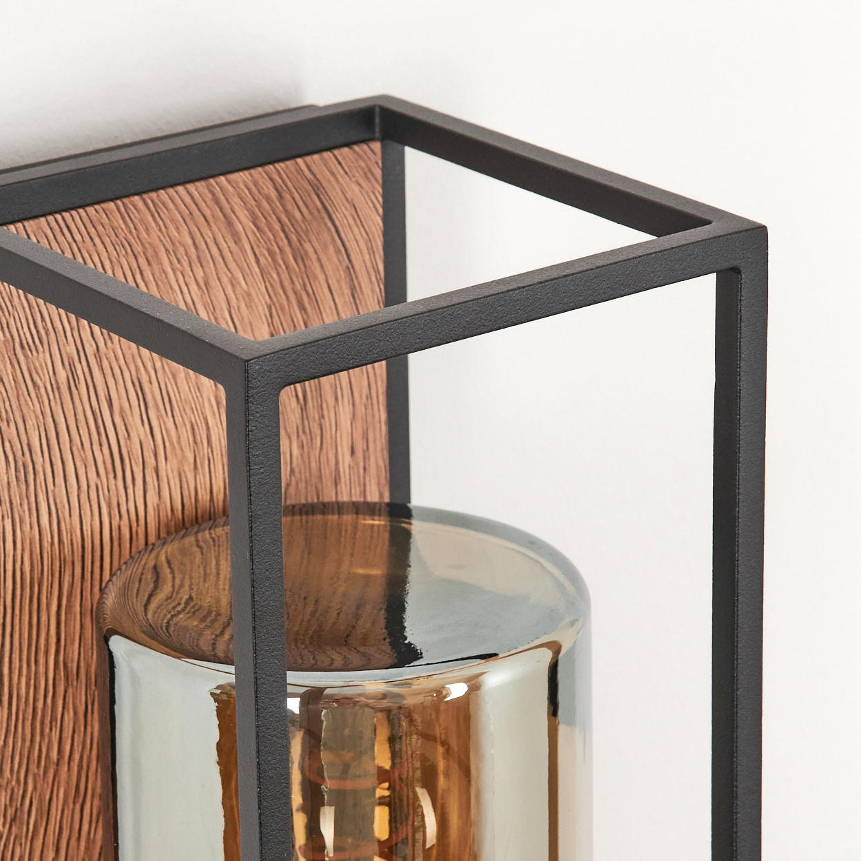 Nahaufnahme einer modernen Lampe mit Holzrücken, schwarzem Rahmen und einem transparenten Glaszylinder mit warmer Glühbirne.