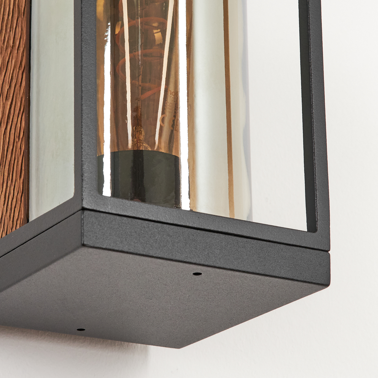 Nahaufnahme einer modernen Lampe mit Holzrücken, schwarzem Rahmen und einem transparenten Glaszylinder mit warmer Glühbirne.