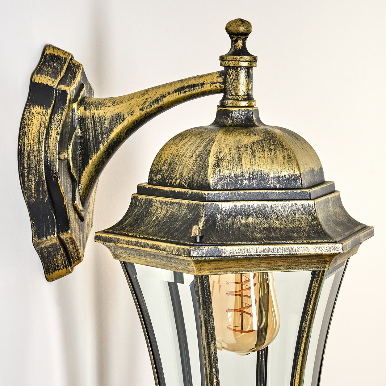 Nahaufnahme der Außenlampe mit Gold- und Schwarz-Finish. Glühbirne sichtbar. An weißer Wand montiert.