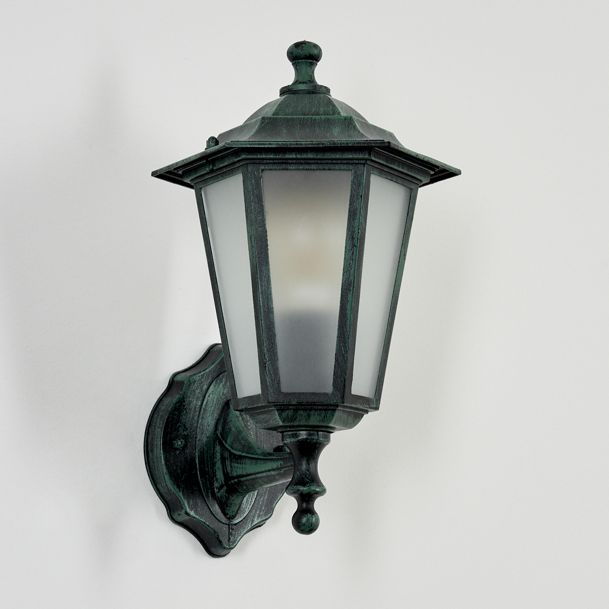 Eine grüne Außenlampe, die an einer weißen Wand montiert ist. Die Lampe hat eine dekorative Oberseite und Milchglas.