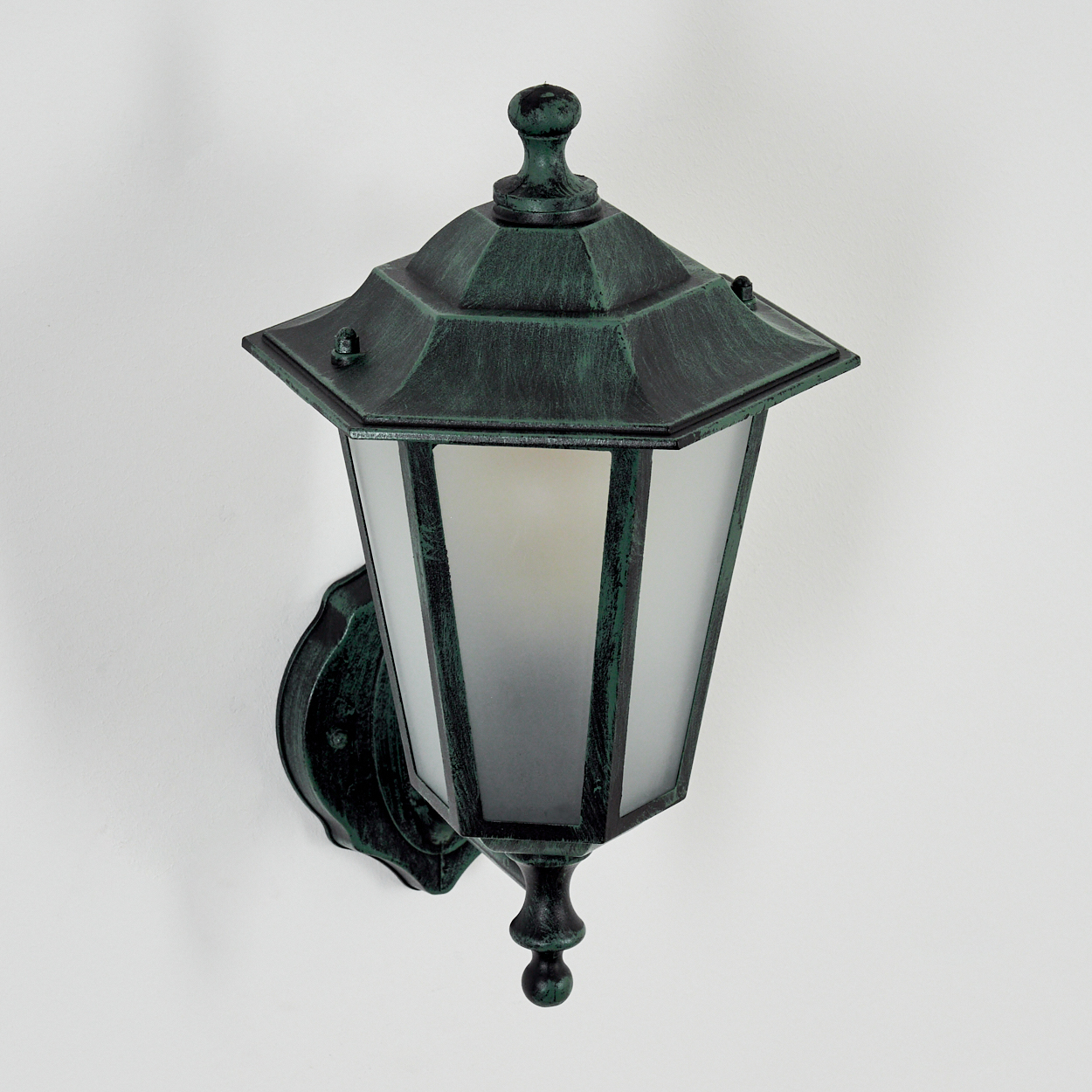 Eine grüne Außenlampe, die an einer weißen Wand montiert ist. Die Lampe hat eine dekorative Oberseite und Milchglas.