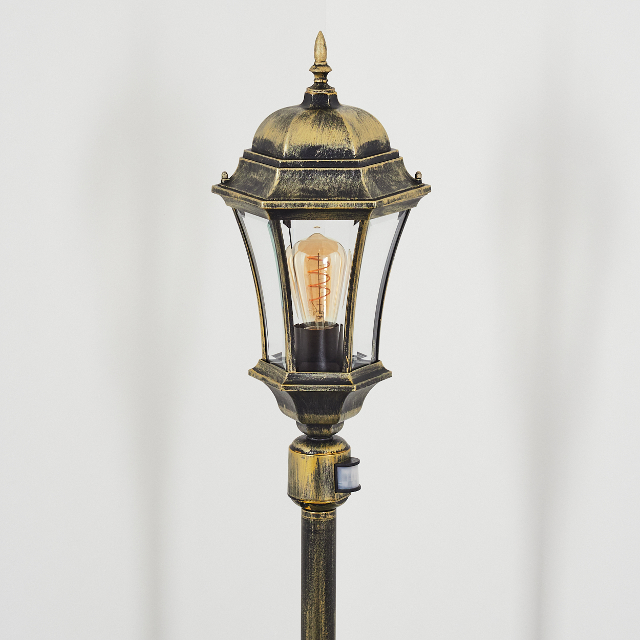 Ein goldener Außenlampenpfosten mit einem Glasgehäuse und einer leuchtenden Glühbirne. Ein Bewegungssensor ist sichtbar. Weißer Hintergrund.