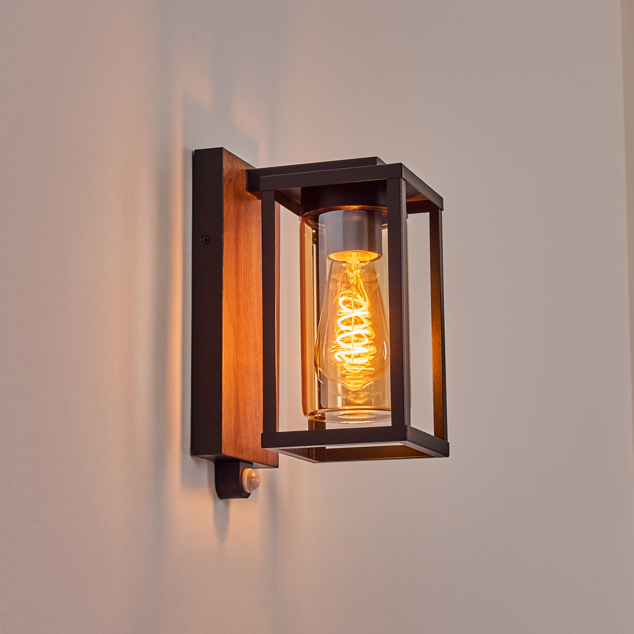 Wandlampe mit Holzsockel, schwarzem Rahmen und einem Zylinder aus Klarglas mit warmtoniger Glühbirne.