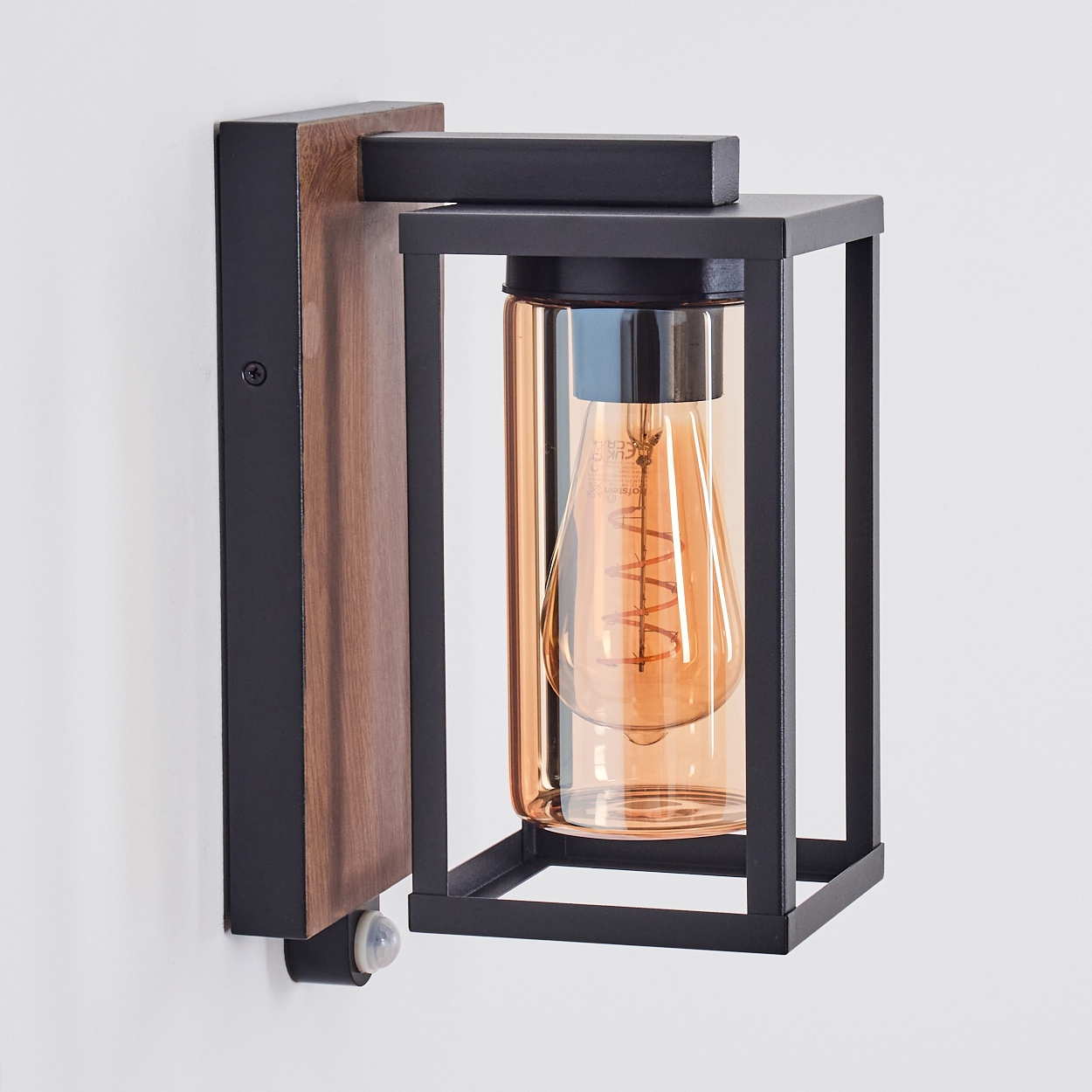Wandlampe mit Holzsockel, schwarzem Rahmen, Klarglas und warmtoniger Glühbirne. An einer weißen Wand.