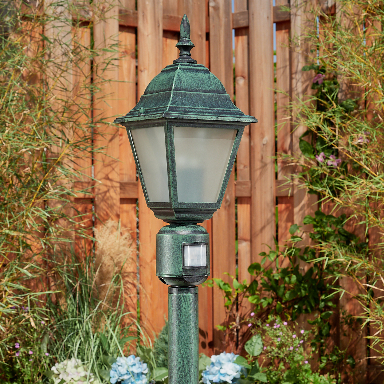 Grüne Laterne mit Lampe, in einem Garten mit Holzzaun und verschiedenen Pflanzen im Hintergrund.
