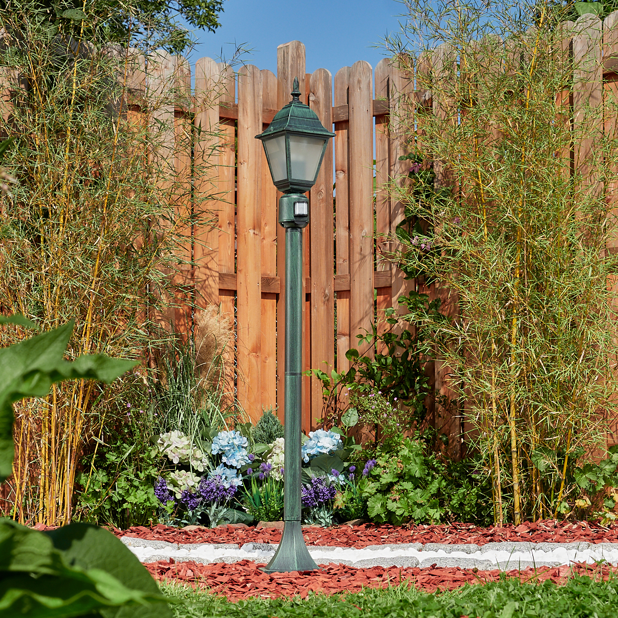 Grüner Laternenpfahl mit Lampe in einem Garten. Holzzaun und Pflanzen im Hintergrund.