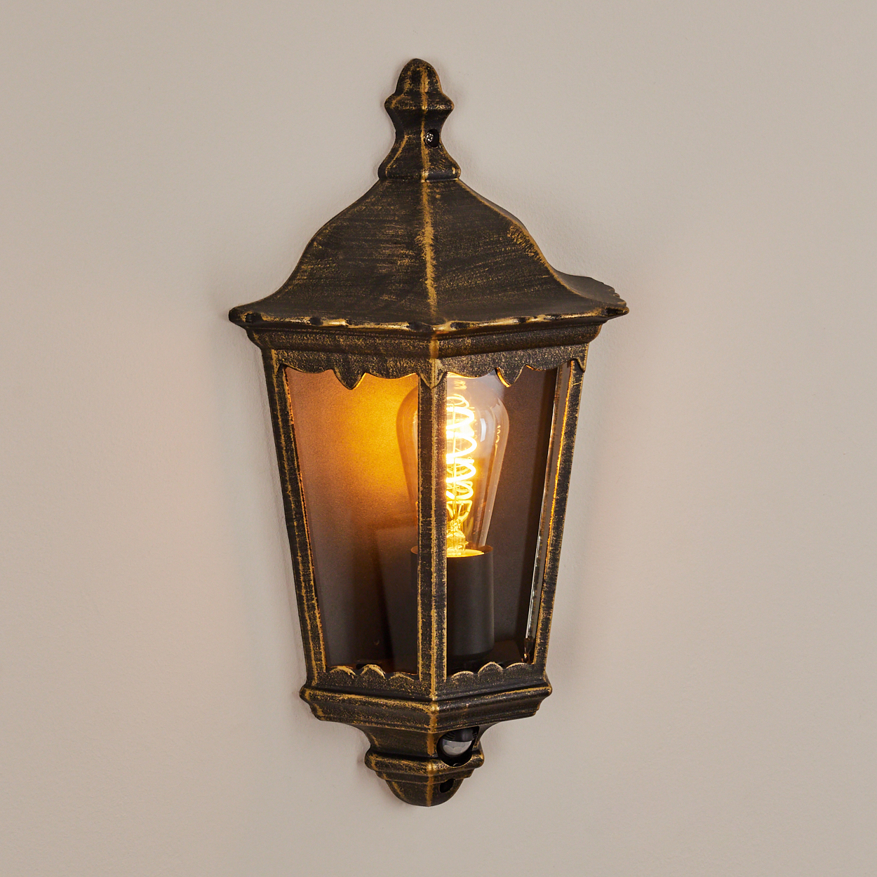 Eine verzierte, goldfarbene Außenwandleuchte mit einer leuchtenden Glühbirne. Die Lampe ist an einer beigen Wand befestigt.