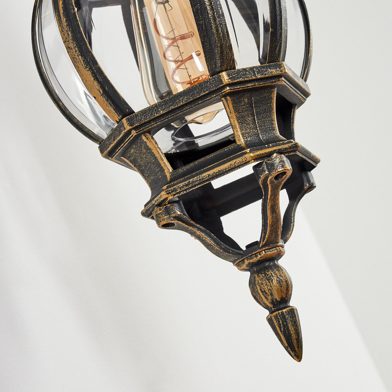 Nahaufnahme einer schwarzen und goldenen Außenlampe mit Glas und einer Glühbirne, vor einer weißen Wand.