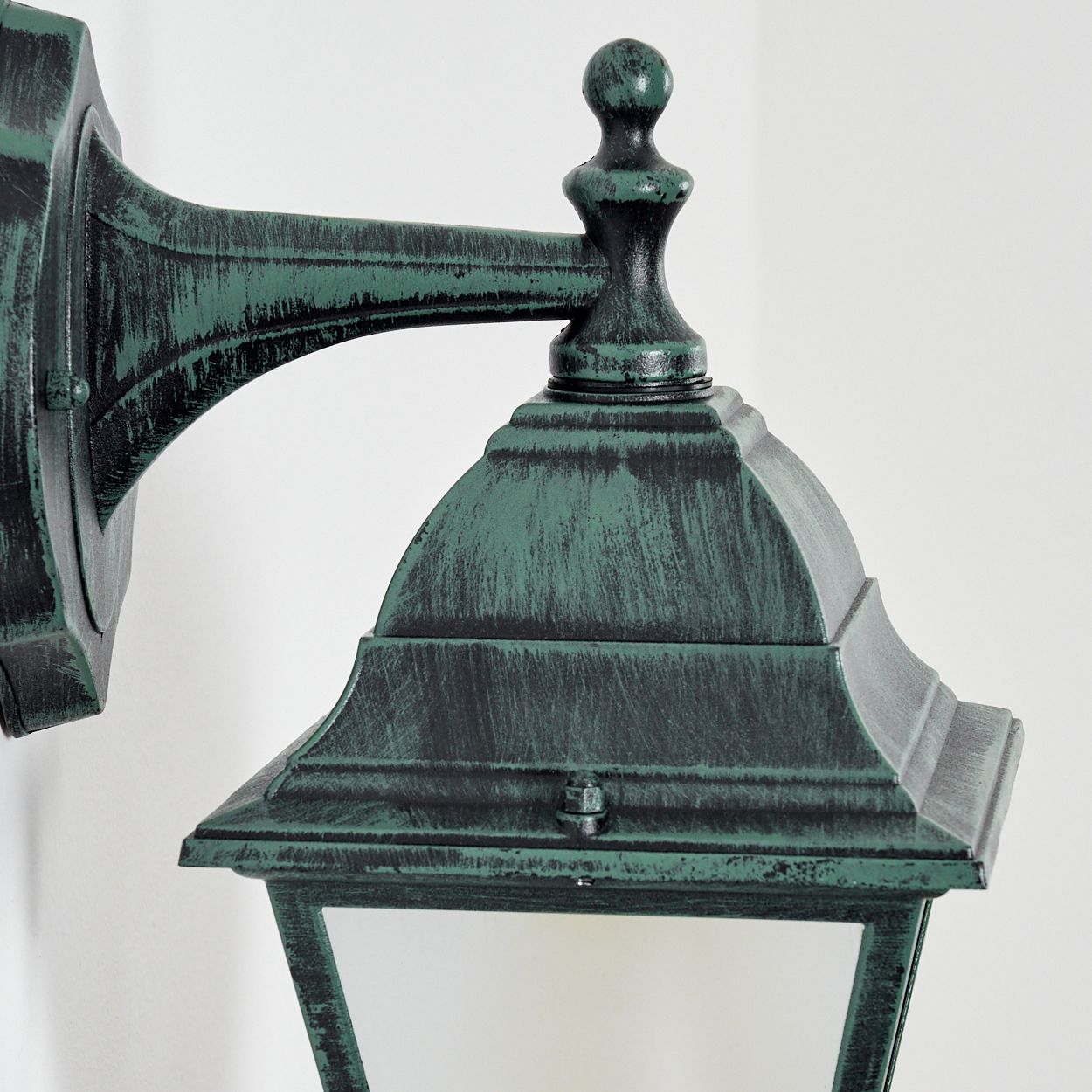 Nahaufnahme einer grünen Außenlampe vor weißer Wand, mit Verzierungen an der Spitze und der Halterung.